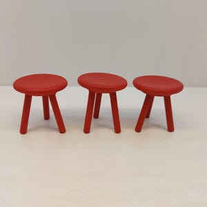 May include: Three small, red wooden stools with rounded seats and three legs each. The stools are evenly spaced against a white background. Each stool is a vibrant shade of red. The stools are a set.