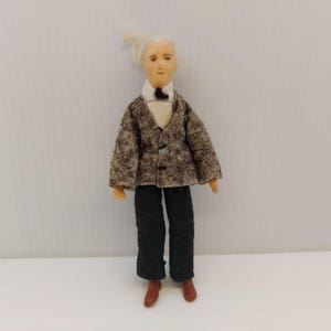 May include: A handmade doll dressed in a miniature suit. The doll has a light complexion, white hair, and is wearing a brown tweed jacket, black pants, a white shirt, and a black bow tie. The doll is wearing brown shoes.