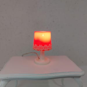 May include: A small, white table lamp with a red and white lampshade. The lampshade has a lace design around the bottom. The lamp is lit, emitting a warm, red glow. The lamp sits on a white table.