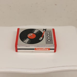 Op de afbeelding: Een miniatuur zilveren en rode speelgoed platenspeler met een zwarte vinylplaat en een rode kern. Het woord "Lundby" staat op de voorkant gedrukt. De platenspeler staat op een wit oppervlak.