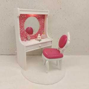 Puede incluir: Un tocador blanco en miniatura con un fondo rosa con estampado floral, un espejo redondo y un cajón pequeño. El conjunto incluye una silla rosa a juego. Los accesorios incluyen frascos de perfume y un peine pequeño.