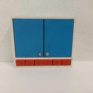 May include: A white cabinet with two blue doors and six red drawers. The cabinet has silver knobs on the doors.