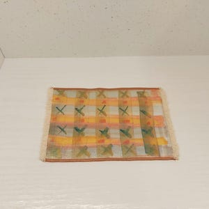 Può includere: Un piccolo tappeto rettangolare con un motivo geometrico. Il tappeto ha uno sfondo bianco con strisce gialle, arancioni e verdi e forme a X. Il tappeto ha un bordo a frangia.