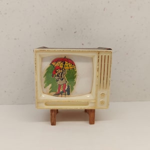 May include: A vintage toy television set with a white plastic body and brown wooden legs. The screen shows a cartoon of a person holding an umbrella.
