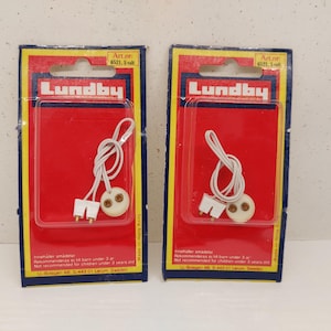 Puede incluir: Dos juegos de luces para casas de muñecas Lundby. Cada juego incluye un cable blanco con una bombilla y un enchufe de dos clavijas, empaquetados en una tarjeta roja con el logotipo de Lundby. El embalaje es rojo, amarillo y azul.