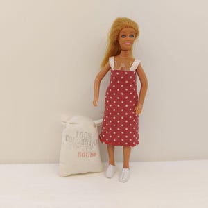 Könnte beinhalten: Eine Puppe in einem roten Polka-Dot-Kleid und weißen Schuhen steht neben einem kleinen Jutesack. Auf dem Sack steht "100% Colombian Coffee 50lbs" in verblasstem Grau und Rot. Die Puppe hat blondes Haar und steht vor einem weißen Hintergrund.