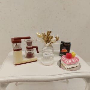 Op de afbeelding: Miniatuur koffiezetapparaat, koffiekopje en cake op een witte tafel. Er staat ook een vaasje met gouden en witte decoratieve stelen. Een klein doosje espresso staat naast de cake.