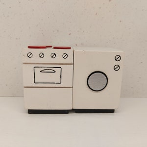 May include: White toy kitchen appliances, a stove and a washing machine, with red knobs on the stove and black markings on both appliances.