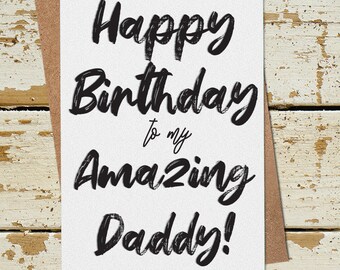 May include: A white card with black text that reads "Happy Birthday to my Amazing Daddy!" The card is on a brown background with a wooden surface.