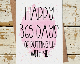 Op de afbeelding: Een roze en witte kaart met de tekst "Happy 365 Days of Putting Up With Me" in zwarte letters. De kaart ligt op een houten oppervlak.