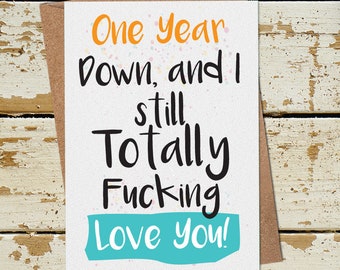 Op de afbeelding: Een wenskaart met de tekst "One Year Down, and I still Totally Fucking Love You!" in zwart, oranje en turquoise. De kaart ligt op een bruine envelop, met een kopje koffie rechtsboven.