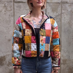 May include: A long-sleeve quilted jacket featuring a multi-coloured patchwork design with various floral and geometric patterns. The jacket has a collared neckline, a black trim along the edges, and a front button closure with five round wooden buttons.