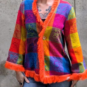 Op de afbeelding: Een jas met lange mouwen en een levendig patchworkontwerp van vierkanten in de kleuren rood, oranje, geel, blauw en paars. Het kledingstuk heeft een enkele knoopsluiting en is afgewerkt met feloranje franjes langs de kraag en mouwen.