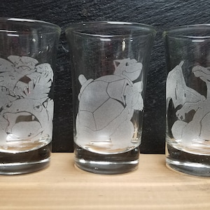 May include: Three clear glass shot glasses with etched designs of Pokemon characters. The first glass features a Bulbasaur, the second glass features a Squirtle, and the third glass features a Charizard.