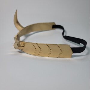 May include: A gold-colored metal crown with a curved horn and chevron design. The crown has a black elastic band for securing it to the head. The design is reminiscent of a horned helmet.