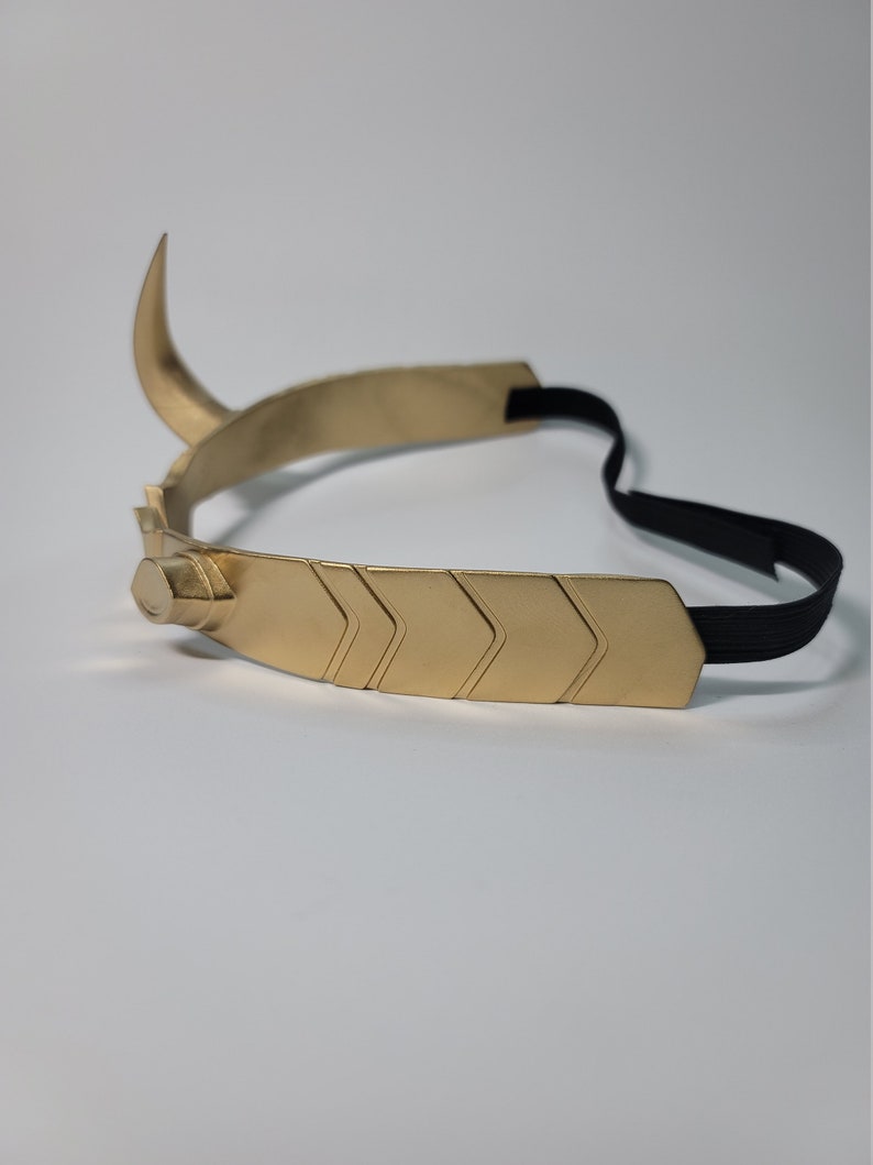 May include: A gold-colored metal crown with a curved horn and chevron design. The crown has a black elastic band for securing it to the head. The design is reminiscent of a horned helmet.
