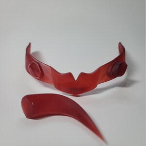 May include: A translucent red plastic accessory, possibly a mask or decorative piece, with a curved band and a separate, pointed element. The band has a central V-shape and two rounded protrusions. The pointed element is curved and tapers to a sharp point.