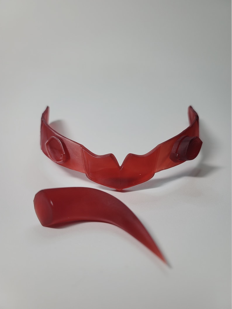 May include: A translucent red plastic accessory, possibly a mask or decorative piece, with a curved band and a separate, pointed element. The band has a central V-shape and two rounded protrusions. The pointed element is curved and tapers to a sharp point.