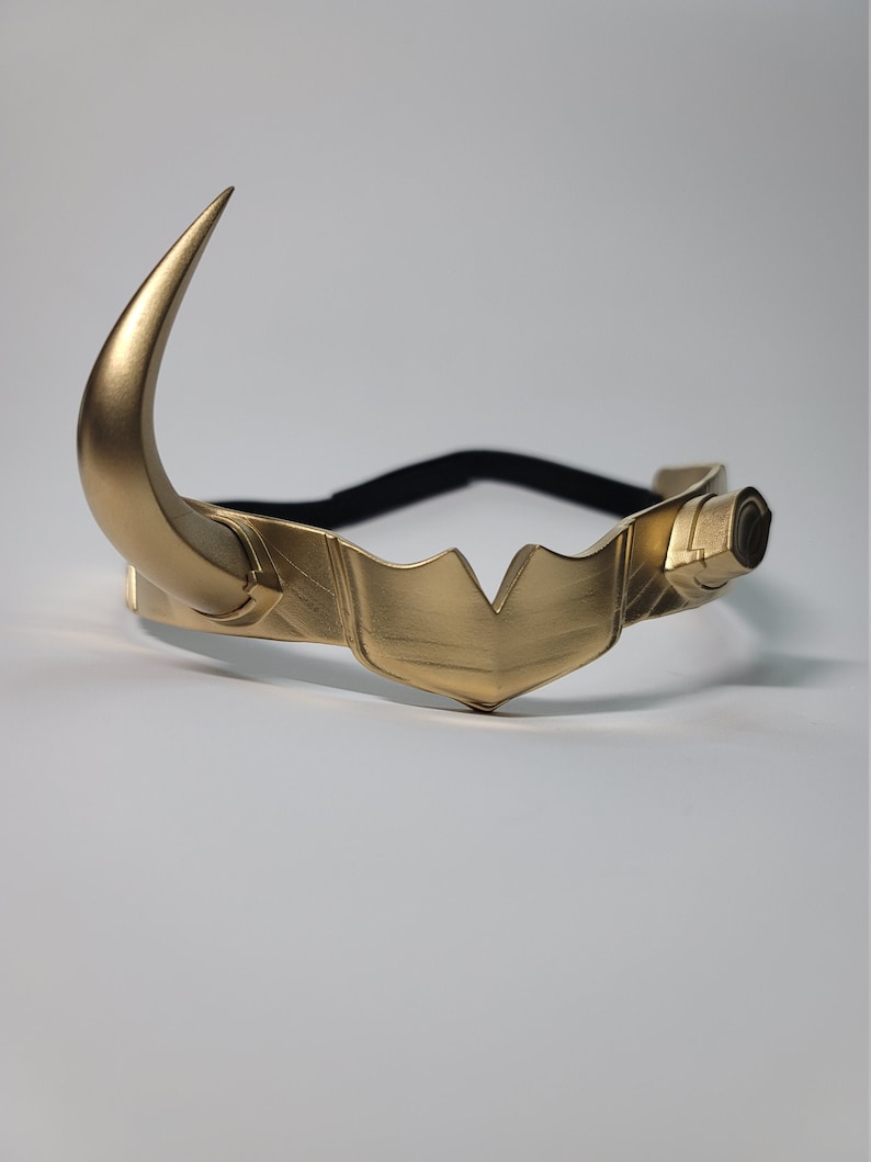 May include: A gold-colored headpiece with two curved horns and a central crest. The headpiece has a black elastic band for wear. The design is metallic and appears to be a costume accessory.