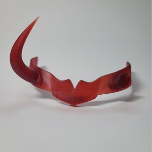 May include: A translucent red headpiece with a curved horn on one side. The headpiece has a central point and rounded details on the sides. The object is set against a white background.