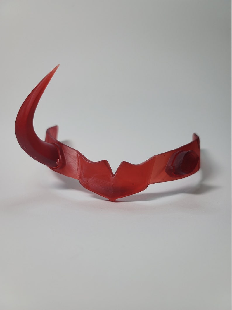 May include: A translucent red headpiece with a curved horn on one side. The headpiece has a central point and rounded details on the sides. The object is set against a white background.