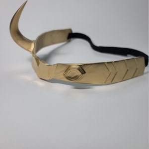 May include: A gold-colored metal crown with a curved horn on the left side. The crown has a central design with an eye-like detail and chevron patterns. A black elastic band is attached for wear.