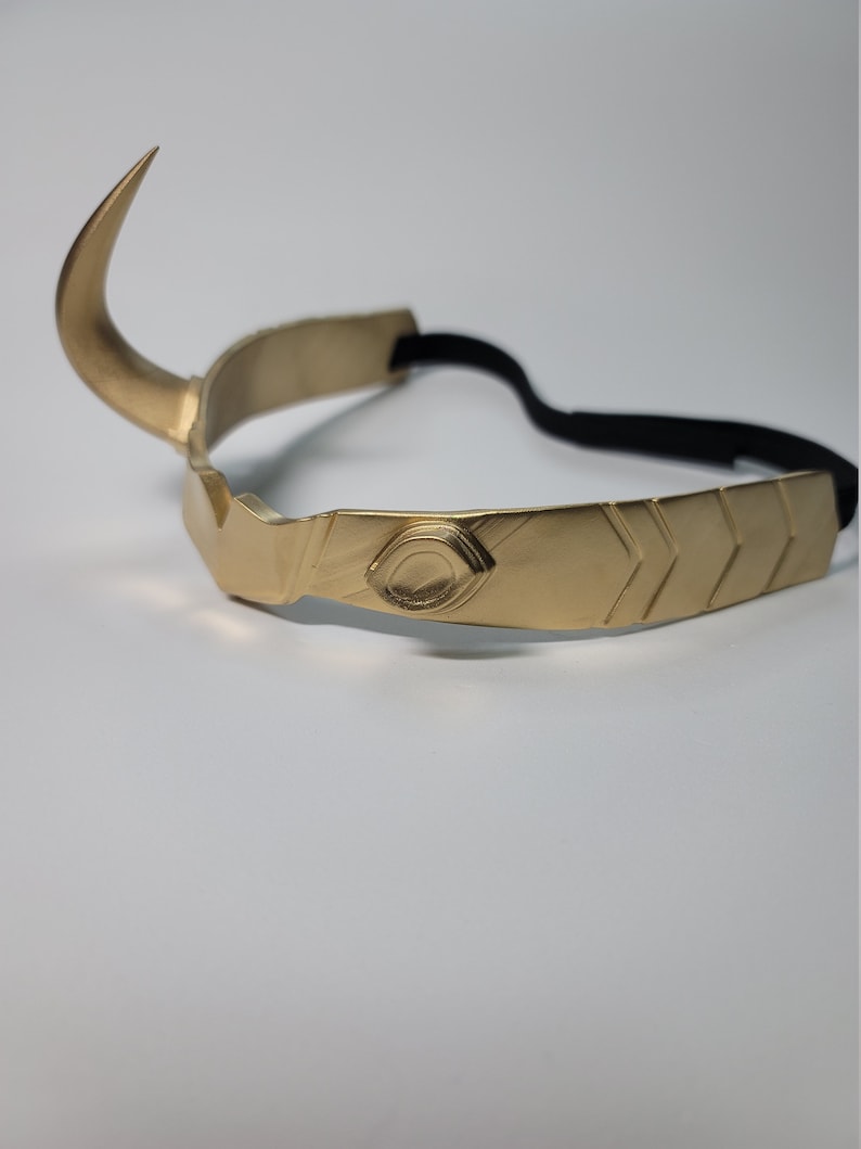 May include: A gold-colored metal crown with a curved horn on the left side. The crown has a central design with an eye-like detail and chevron patterns. A black elastic band is attached for wear.