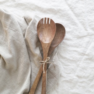 Puede incluir: Juego de servir ensalada de madera. El juego incluye una cuchara y un tenedor con cabezal ranurado. Los utensilios están atados con hilo y descansan sobre un paño de lino beige.