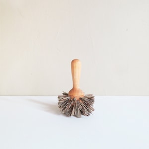 Puede incluir: Un cepillo de madera con cerdas naturales. El cepillo tiene un mango de madera y una base redonda con cerdas que se extienden en todas direcciones.