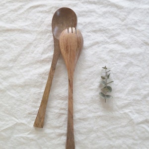 May include: Two wooden salad servers, a spoon and a fork, rest on a white linen surface. The spoon has a rounded bowl and long handle. The fork has a slotted head. A sprig of green leaves is next to the servers.