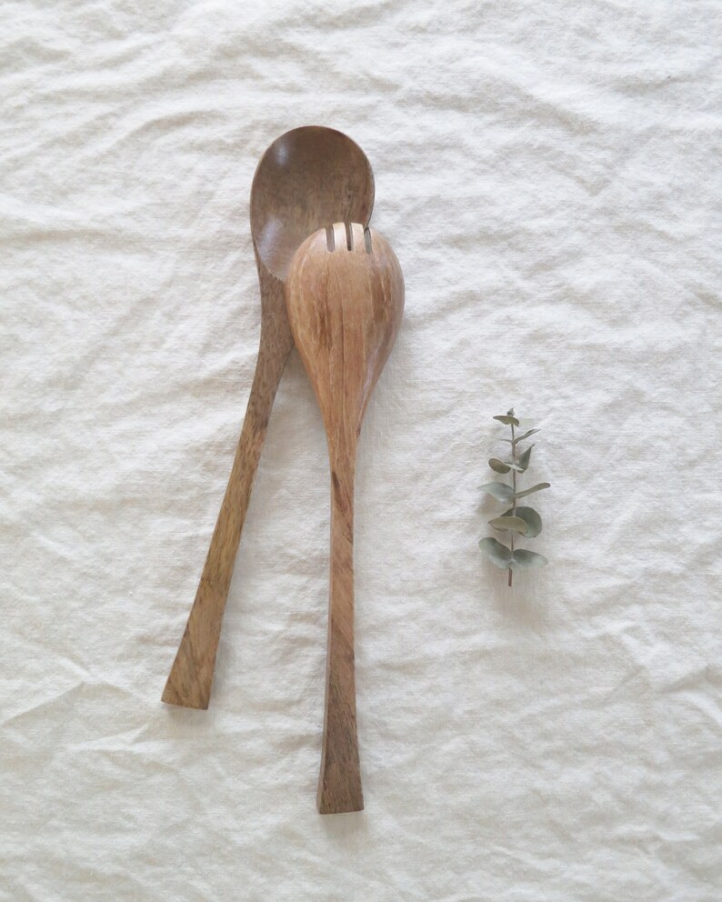 May include: Two wooden salad servers, a spoon and a fork, rest on a white linen surface. The spoon has a rounded bowl and long handle. The fork has a slotted head. A sprig of green leaves is next to the servers.