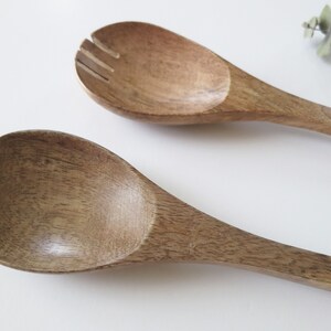 May include: Two wooden salad servers. One server is a solid spoon, and the other has a slotted design. The servers are made of a light brown wood with visible grain. The spoons are on a white background.