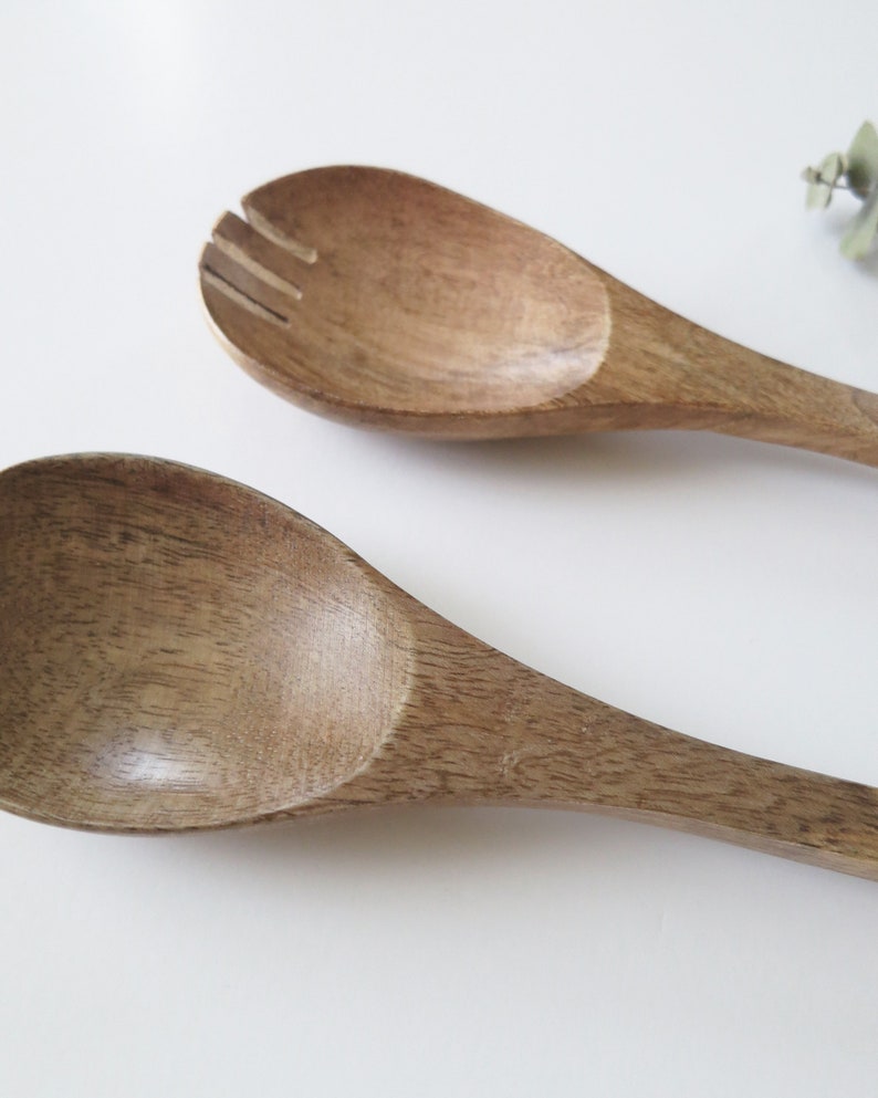 May include: Two wooden salad servers. One server is a solid spoon, and the other has a slotted design. The servers are made of a light brown wood with visible grain. The spoons are on a white background.