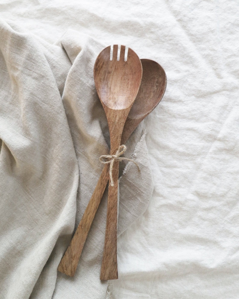 May include: Wooden salad serving set. The set includes a spoon and a fork with a slotted head. The utensils are tied together with twine and rest on a beige linen cloth.