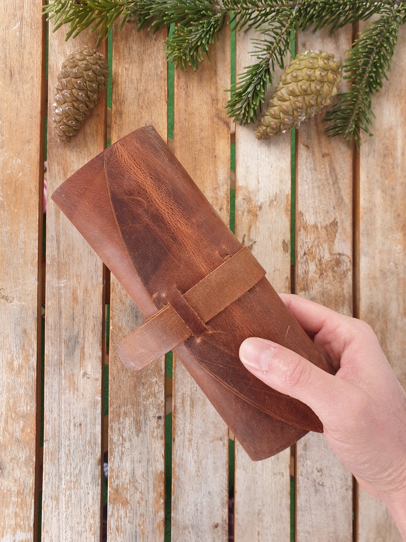 May include: A brown leather roll-up case with a strap closure. The case is made of distressed leather and has a rustic look.