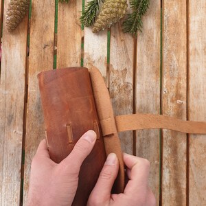 May include: A brown leather pouch with a strap closure. The pouch is being held by two hands.