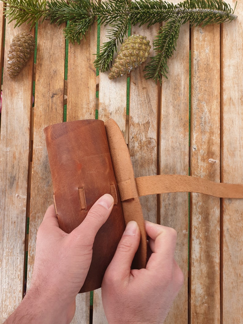 May include: A brown leather pouch with a strap closure. The pouch is being held by two hands.