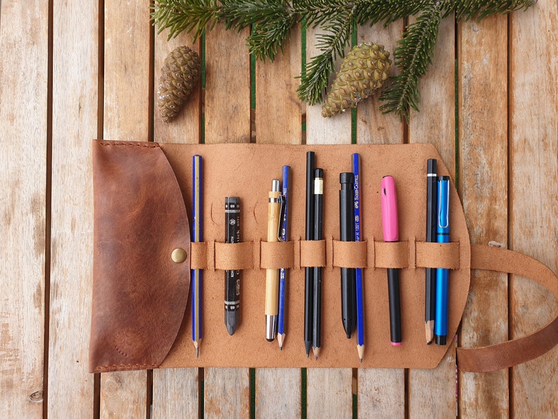 May include: A brown leather pencil roll with multiple slots for holding pencils, pens, and other art supplies. The roll is unrolled and laid flat on a wooden surface.