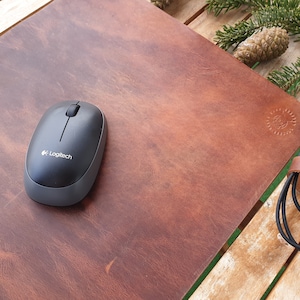 May include: A brown leather mouse pad with a black computer mouse on top. The mouse pad has a small logo embossed in the lower right corner. The mouse is a wireless Logitech mouse.