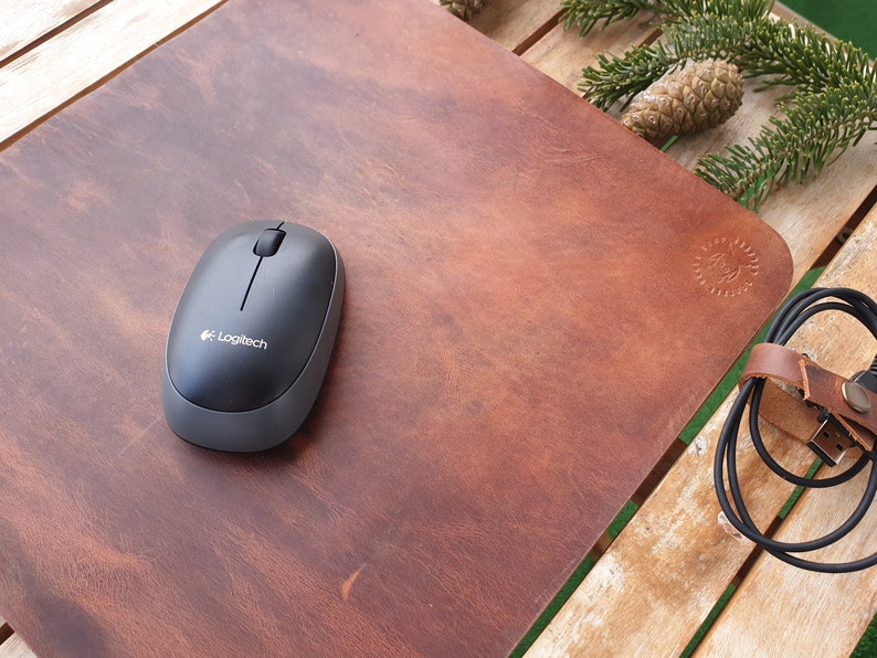 May include: A brown leather mouse pad with a black computer mouse on top. The mouse pad has a small logo embossed in the lower right corner. The mouse is a wireless Logitech mouse.
