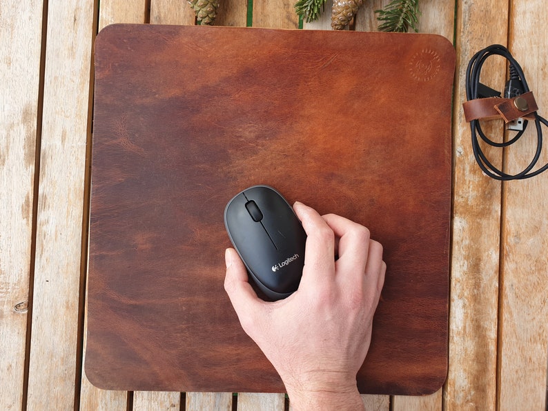 May include: A brown leather mouse pad with a black computer mouse on top. The mouse has the Logitech logo on it.