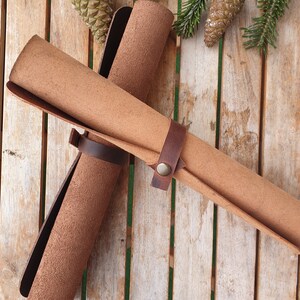 May include: Two brown leather rolls are tied together with a brown leather strap and a metal buckle. The rolls are on a wooden surface with pine cones.