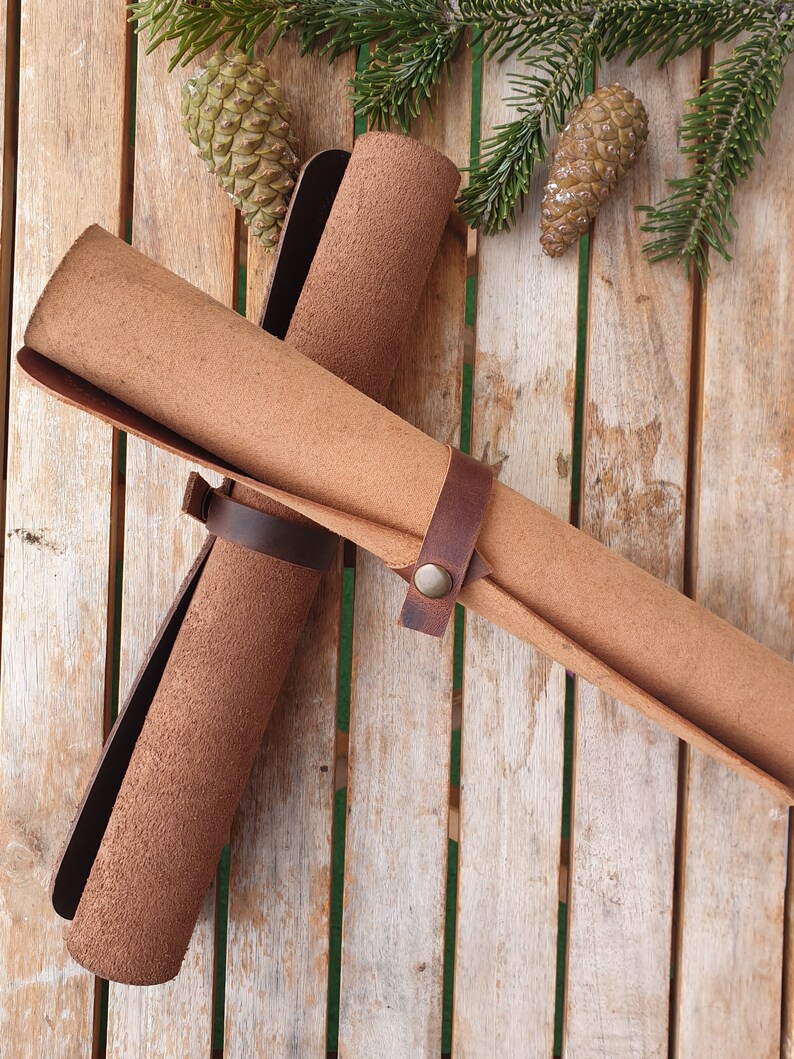 May include: Two brown leather rolls are tied together with a brown leather strap and a metal buckle. The rolls are on a wooden surface with pine cones.