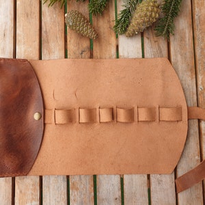 May include: A brown leather roll-up case with a flap closure and multiple slots for holding tools or pens. The case is made of high-quality leather and has a rustic look.