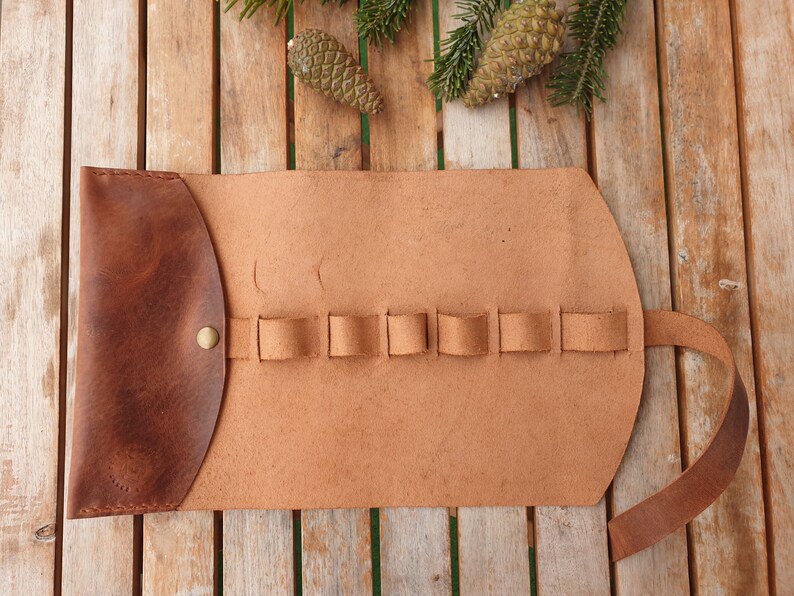 May include: A brown leather roll-up case with a flap closure and multiple slots for holding tools or pens. The case is made of high-quality leather and has a rustic look.