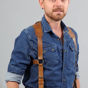 Puede incluir: Un hombre con una camisa vaquera azul y una correa de cámara de cuero marrón. La correa tiene una hebilla y un anillo en la correa del hombro. El hombre lleva un cinturón de cuero marrón.