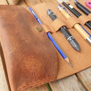 May include: A brown leather pencil roll with multiple slots for pencils and pens. The roll has a brass button closure and a logo embossed on the front.