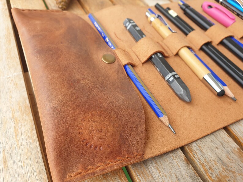 May include: A brown leather pencil roll with multiple slots for pencils and pens. The roll has a brass button closure and a logo embossed on the front.