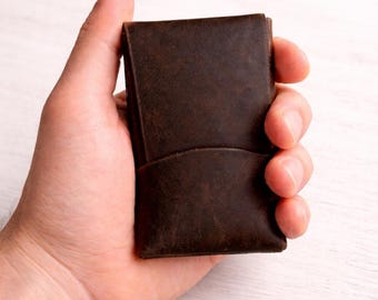 May include: A dark brown leather card holder with a smooth, matte finish and a curved pocket design on the front. The rectangular accessory features visible stitching along the edges and a deep, rich tone with subtle natural grain texture.