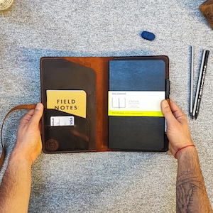 May include: A brown leather notebook cover with a strap, a pocket for a card, and a pen holder. The cover is open to reveal a black notebook with the text "Field Notes" on the front.